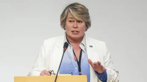 PA Media A woman with blonde hair, wearing a white suit and blue shirt, speaks at a podium in front of a grey background 