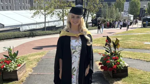 Keele University A woman with long blonde hair is wearing black graduation clothing and a white dress including floral designs. She is standing on a walkway with grass either side of it. A grey building and people are in the distance.