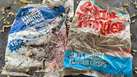 Neil Collett Two Smiths crisp packets from 1975 laid side by side, a blue and white one on the left and the one on the right labelled Golden Wonder in red writing.