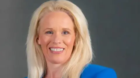 NIAO/PA Comptroller General and Auditor, Dorinnia Carville, looking at the camera. She is wearing a blue jacket with a white necklace. She has blonde hair and is smiling.