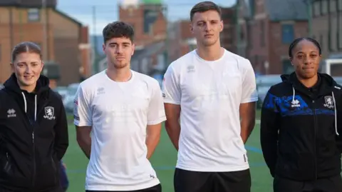Middlesbrough FC Middlesbrough players Hayden Hackney and Dael Fry wearing an all-white football shirt. They're standing between two women in black Middlesbrough tracksuits.