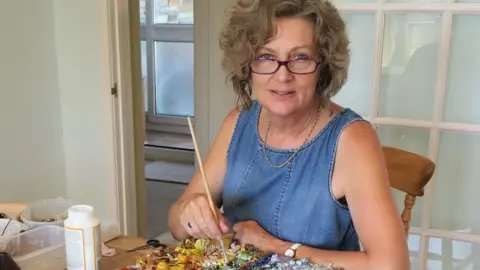 Jane Perkins An artist holds a paintbrush over a collage of tiny plastic items and a pot of glue. She's making a brightly coloured piece inspired by the painter van Gogh