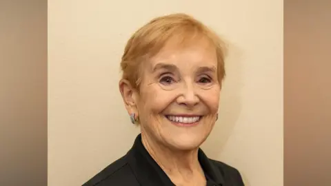 An elderly woman is smiling. She has short-cropped strawberry blonde hair and is wearing earings and eyeshadow. 