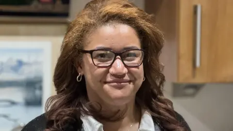 Jamaica is standing in Don's kitchen with a head height brown wall cupboard over her left shoulder. Smiling broadly she's wearing a black and white cardigan over a white shirt. A gold pendant with the letter 'J' on it sits round her neck. 