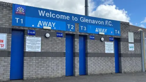 BBC A picture taken outside Glenavon Football Club in Lurgan. There is a 'Welcome to Glenavon FC' sign, with four ticket booths within the grey brick wall. There is a metal gate to the right of the image.
