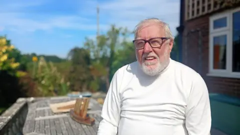  Ian Brittain, wearing a white top and glasses, squints in the sun as he talks in his garden