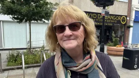 A woman with shoulder length hair and sunglasses, standing in Bangor town centre. It is a head and shoulders shot and she is looking at the camera. She has a brown jumper on and a scarf in an array of dark colours.