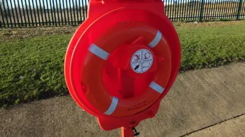 Northamptonshire Search and Rescue A circular red plastic cabinet on a pole, installed on a concrete path by a grass bank. The hinged lid is open and a red lifebuoy is visible inside, attached via white velcro.