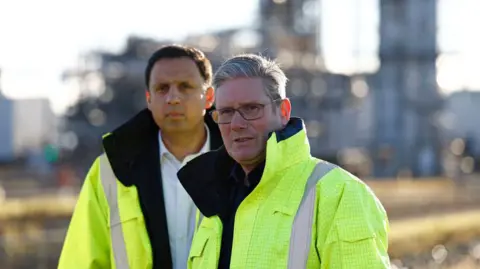 AFP via Getty Images Keir Starmer, who has grey hair and glasses, and Anas Sarwar, who has short black hair, are both wearing high-vis yellow jackets as they stand outside an industrial complex
