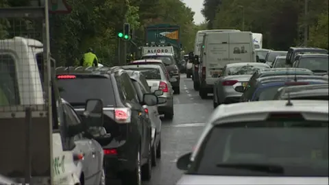 Three lanes of back-to-back traffic heading away from the camera, with green trees wither side of the road.