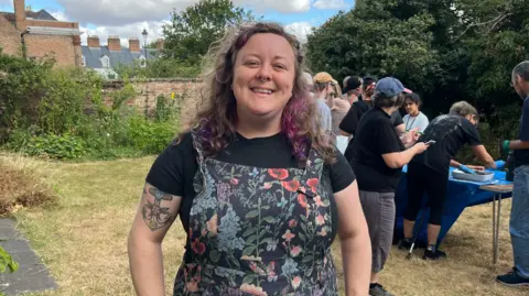 Programme organiser Lizzy standing in front of the gardening group working on their project. She is wearing floral dungarees with a black T-shirt underneath. Her hair is brown with purple streaks and she has a septum piercing and a tattoo on her left upper arm.