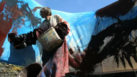 Tszman Chan A person wearing a paint‑splattered garment stands outdoors holding a container and a cup coated in paint.
Behind the person, a large sheet covered in broad, abstract strokes of blue, black, and red paint stretches across the scene.