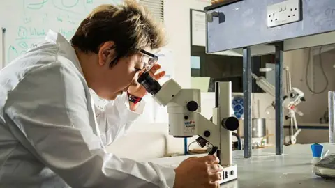 Asel Sartbaeva sits in a lab and looks through a microscope. She has short brown hair with highlights. She wears a white lab coat and has rimless glasses resting above her eyes.