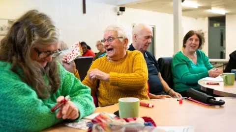 Damien Wooten A number of people playing bingo. One lady laughs as she holds up her card and shows it to someone. She has grey hair and is wearing a mustard jumper and black-rimmed glasses. She is sitting next to a woman with longer grey hair who is wearing a green jumper. 