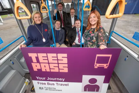 Tees Valley Combined Authority A group of six individuals, four men and two women, are a mixture of sitting and standing on a bus. In front of the group is a poster being held up with the text "Tees Pass" "Your Journey Free Bus Travel".