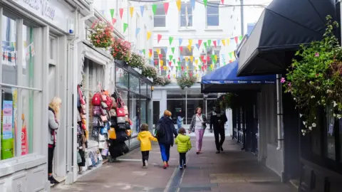 A paved street in St Peter Port lined with shops. There is a woman holding the hands of two children walking down it and a man and a woman walking up it. A lady is standing in a shop door way. There is mulit-coloured bunting strung across the lane and one of the shops has bags hung outside it.