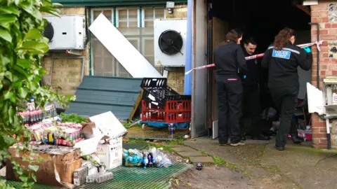 Martin Giles/BBC The back of residential and shop buildings. Several cases of Cola can be seen outside the building. A cordon prevents entry into the back of the store. Three police staff can be seen. 