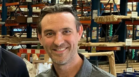 Kevin Utton smiling at the camera. He is stood in front of shelves stacked with objects. He has short dark hair and stubble, and has sunglasses on the top of his head.