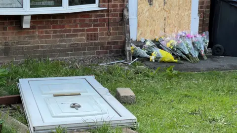 Tom Ingall/BBC Several bunches of flowers left on the doorstep of a house. The door has been replaced by a wooden board and the door to the house is lying on the ground in the front garden.