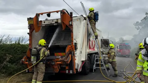 Fire fighters work on a white bin lorry, with one spraying water into the rear and another on a ladder inspecting the top.