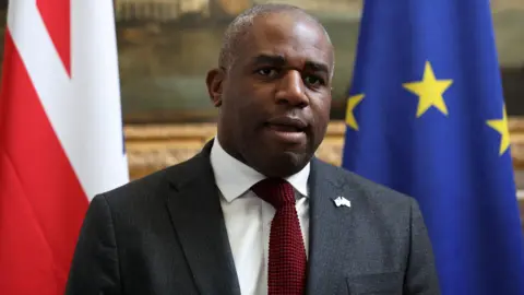 British Foreign Minister David Lammy wearing a suit and red tied speaks to reporters stood in front of a union flag and a flag of the European union ahead of meeting European Union High Representative for Foreign Affairs and Security Policy Kaja Kallas (not pictured)