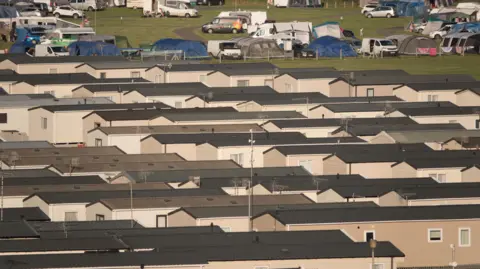 The roofs of multiple rows of static caravans stretch out across a field. Beyond the caravans, there are tents and parked vehicles dotted around the site.