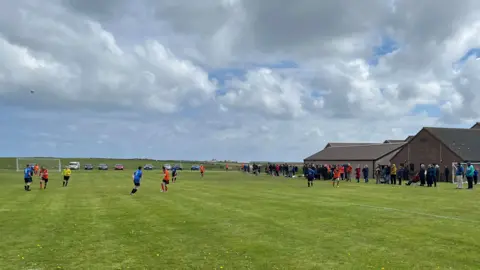 Men in orange and black playing football. Supporters watching at the side of the pitch.