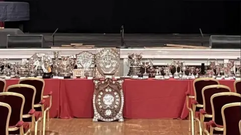 MANX MUSIC FESTIVAL A table of trophies, with a large shield in the foreground and chairs on either side, there is a stage in the background.