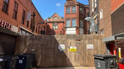 An alleyway, with buildings and bins either side, which has been blocked off by a wooden fence with warning signs attached to it.