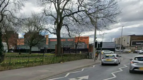 Google Two silver approach a roundabout on Ardwick Green South with a park on the right and the Apollo music venue ahead