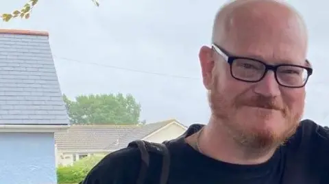 Ian Airlie is seen with glasses and a beard is smiling outdoors, with houses and trees visible in the background on a cloudy day.