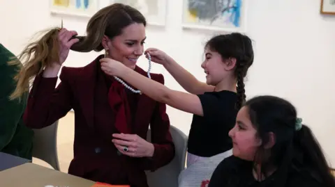 A girl holds up a necklace around the Princess of Wales' neck, while another girl looks on
