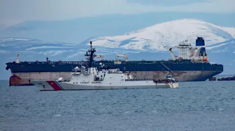 Peter Jolly/Northpix The tanker is a large ship with rusty marks on its hull. The upper part of its hull is painted blue and it has a white bridge and a single blue funnel. In the foreground is a white US Coast Guard vessel. There are snow-covered hills in the background.
