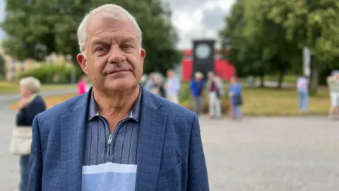 Sir Mark is standing outdoors, wearing a blue blazer over a striped shirt. In the background, several people are gathered in a park-like setting with trees and a large clock structure visible. The sky is partly cloudy