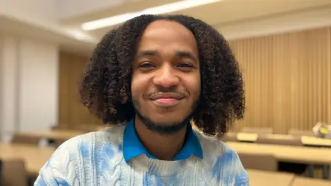 A man with shoulder length hair is looking directly at the camera. He is sat in front of a row of desks, wearing a shirt with a blue collar and a blue and white tie dye patterned cable knit jumper.