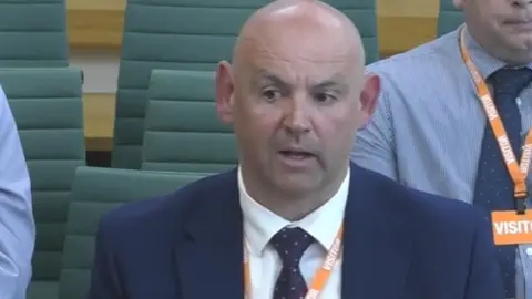 Parliament TV A man sitting down in a green chair with other chairs and two people in the background. He has a bald head and is wearing a blue suit and tie with a white shirt underneath. 