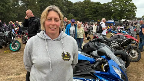 BBC A woman stood in front of a row of motorcycles, smiling at the camera. She has long, blonde hair tied back and is wearing a grey hoodie with a black and yellow circular logo on. There are groups of people admiring the motorcycles behind her. 