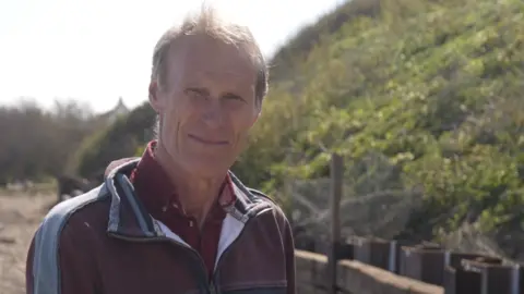 James Crowhurst standing on the beach at Thorpeness. He has short light brown hair and is squinting slightly in the sun, wearing a maroon fleece over a maroon shirt. Behind him is a wooden barrier and behind that sheet metal piling, installed in a zig zag pattern. Rising up from it is a shallow leafy cliff. 