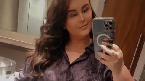 Cathryn Russell A selfie of a young woman with long brown hair wearing a silky grey crinkle shirt. She is holding up a black phone and standing in a bathroom with brown tiled floor and walls and a brown door and mirror behind her.