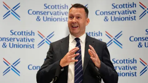 Russell Findlay gestures with his hands astatine  the media league  for his predetermination  arsenic  leader. He is lasting  successful  beforehand   of Scottish Conservative enactment      branding.