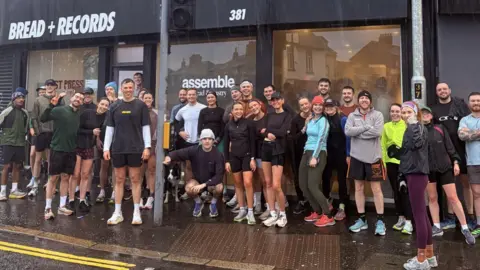 Run East A group of runners is assembled on a footpath outside a local bakery. The shop sign reads Bread and Records. The runners are wearing t-shirts and shorts in various colours 