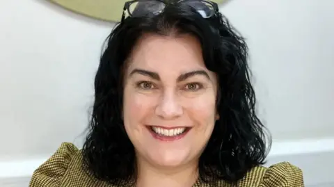 Streetlife Trust Kim Hughes with long dark brown hair wearing sunglasses on her head and a yellow and black checked top. She is smiling.