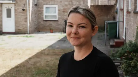 John Fairhall/BBC Woman with her hair tied back smiling at the camera. Behind her is a house and some grass. She is wearing a black top.