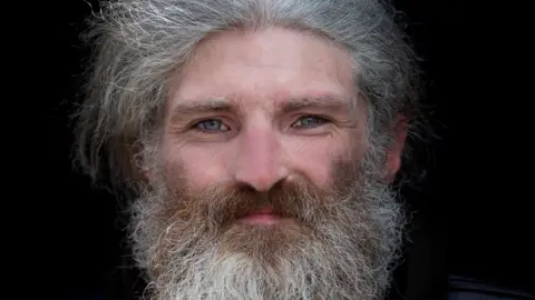 Jennifer Charlton Long grey haired and bearded man with piercing blue eyes looking at the camera.