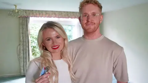 Georgia Pickford and Cameron Smith standing in their living room. She holds the keys to their new home