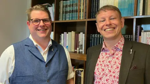 BBC/Fiona Callow Two men stand in front of a full bookshelf, smiling. The man on the left has brown hair combed back and wears black, round glasses and a blue waistcoat over a white short-sleeved shirt. The man on the right has short brown hair and wears a white shirt patterned with red birds and a brown jacket.