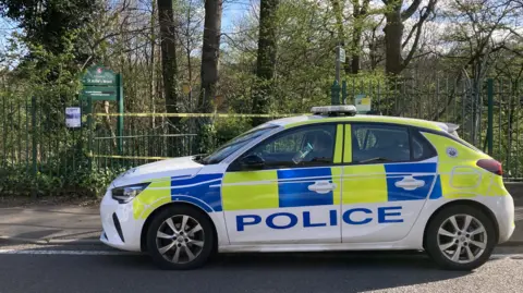 A police car with yellow and blue hi-vis decals parked outside the entrance to St Anne's Wood, alongside the green metal fence. There is a yellow cordon tape securing the gated entrance.