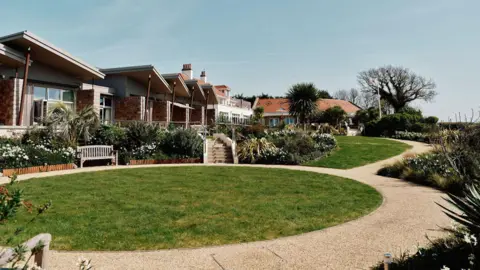 Jersey Hospice Care The hospice has units which are single storey and have balconies. There is circle of lawn with a wooden seat overlooking it and a path around it. There are also flower beds and palm trees.