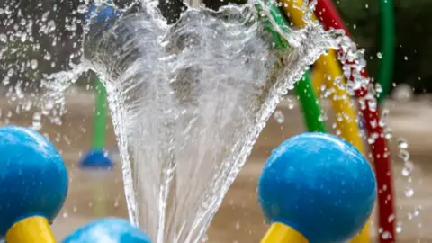 A spout of water heading upwards with droplets falling off either side. There are several colourful items blurred in the background.