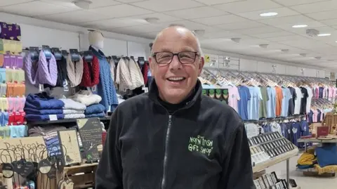 Keith Lane Keith Lane. He is bald, wearing glasses and a black zip-up fleece with green writing saying 'The Norn Irish Gift Shop'. Behind is wracks of t-shirts and scarfs and folded knits.